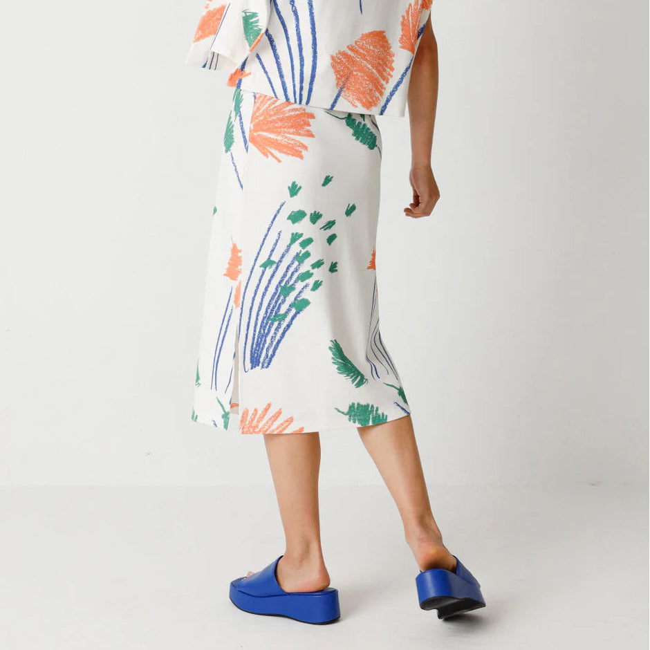 Eine Frau steht mit dem Gesicht zur Wand und trägt ein weißes Outfit mit orangefarbenen, grünen und blauen Blumenmustern. Sie hat schulterlanges braunes Haar und blaue Slipper. Der Hintergrund ist einfarbig weiß.