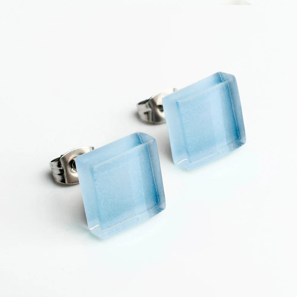 Ein Paar Ohrstecker mit quadratischen, durchscheinenden hellblauen Steinen in Metallfassungen auf weißem Hintergrund.