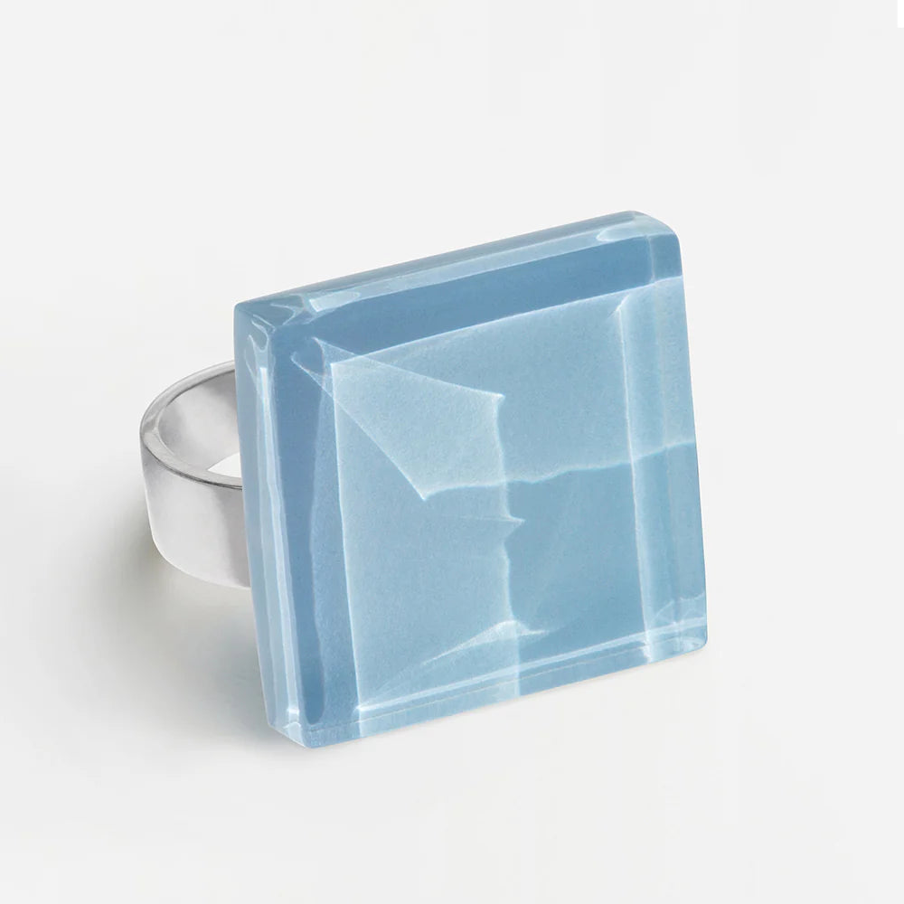 Ein Silberring mit einem großen, quadratischen, durchscheinenden blauen Edelstein mit geometrischer, facettierter Oberfläche auf weißem Grund.