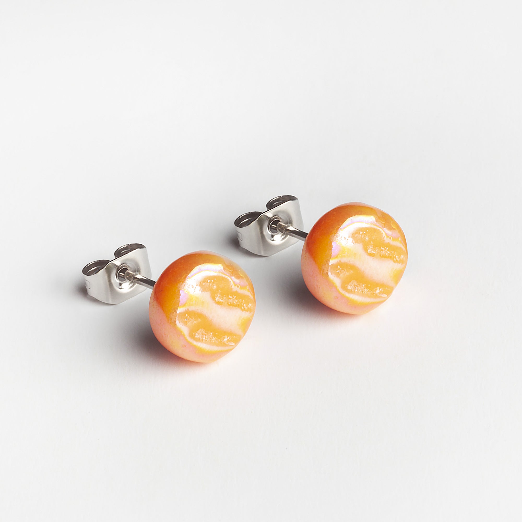 Ein Paar Ohrstecker mit runden, orange-weiß gewirbelten Mustern, die an Zitrusfruchtscheiben erinnern, auf silbernen Stiften vor einem weißen Hintergrund.