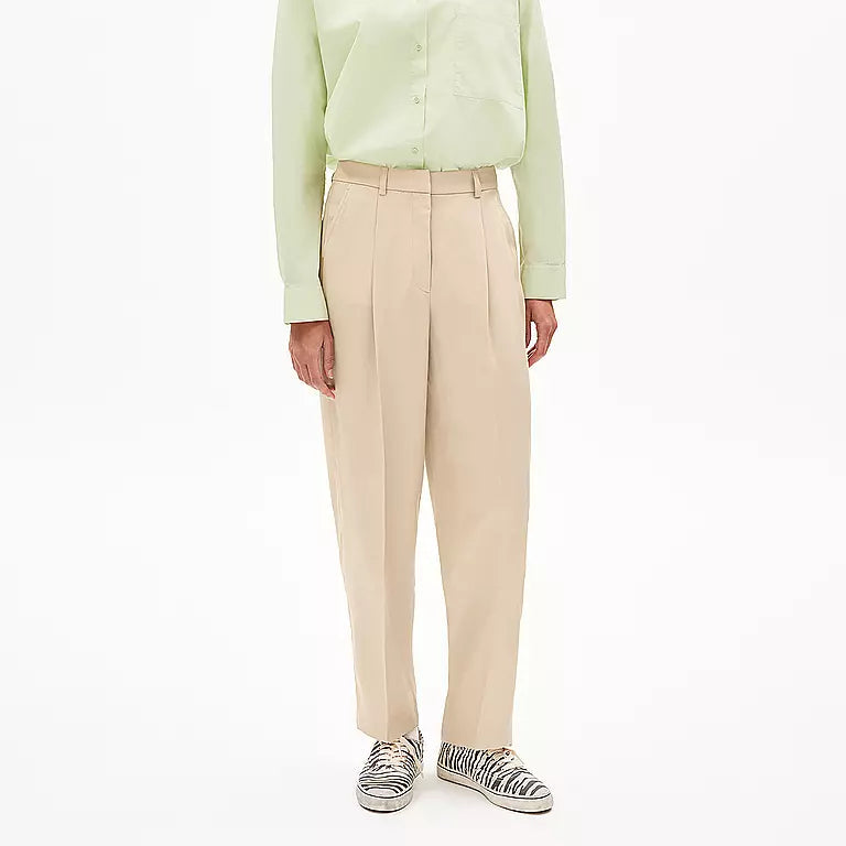 Eine Person, die ein hellgrünes Button-up-Hemd, eine beigefarbene Bundfaltenhose und Turnschuhe mit Zebrastreifen trägt, steht vor einem schlichten weißen Hintergrund.