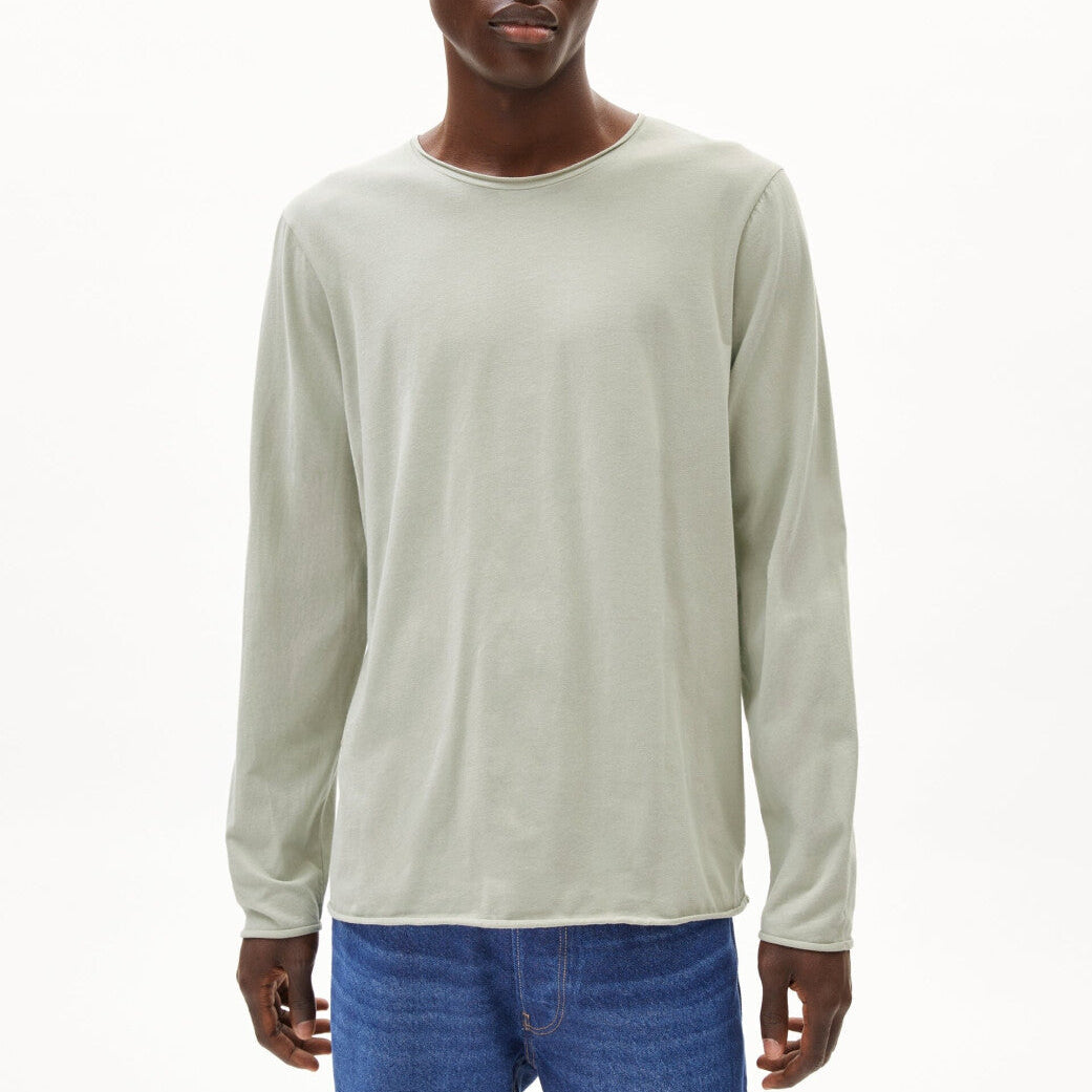 Eine Person, die ein hellgraues langärmeliges Hemd und blaue Jeans trägt, steht vor einem schlichten weißen Hintergrund. Das Hemd hat eine einfache, lockere Passform und einen runden Ausschnitt.