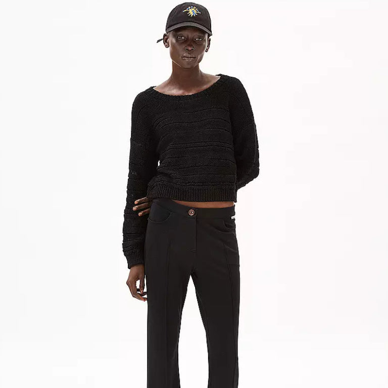 Eine Person, die einen schwarzen Strickpullover, eine schwarze Hose, eine schwarze Baseballmütze mit einem bunten Muster und weiße Schuhe trägt, steht vor einem schlichten weißen Hintergrund.