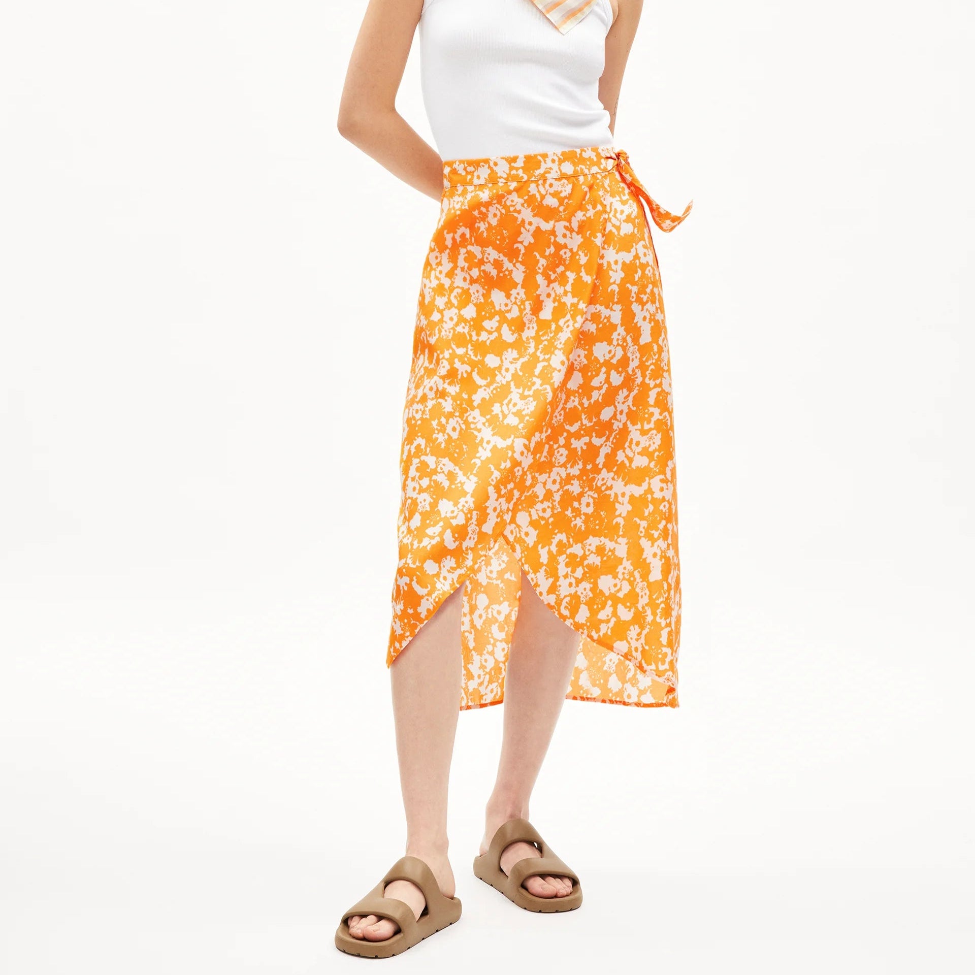 Eine Frau steht vor einem schlichten Hintergrund und trägt ein weißes ärmelloses Oberteil, einen orangefarbenen geblümten Wickelrock, einen gestreiften Schal um den Hals und braune Sandalen. Ihre Arme sind entspannt, und ihr dunkles Haar ist glatt und locker.