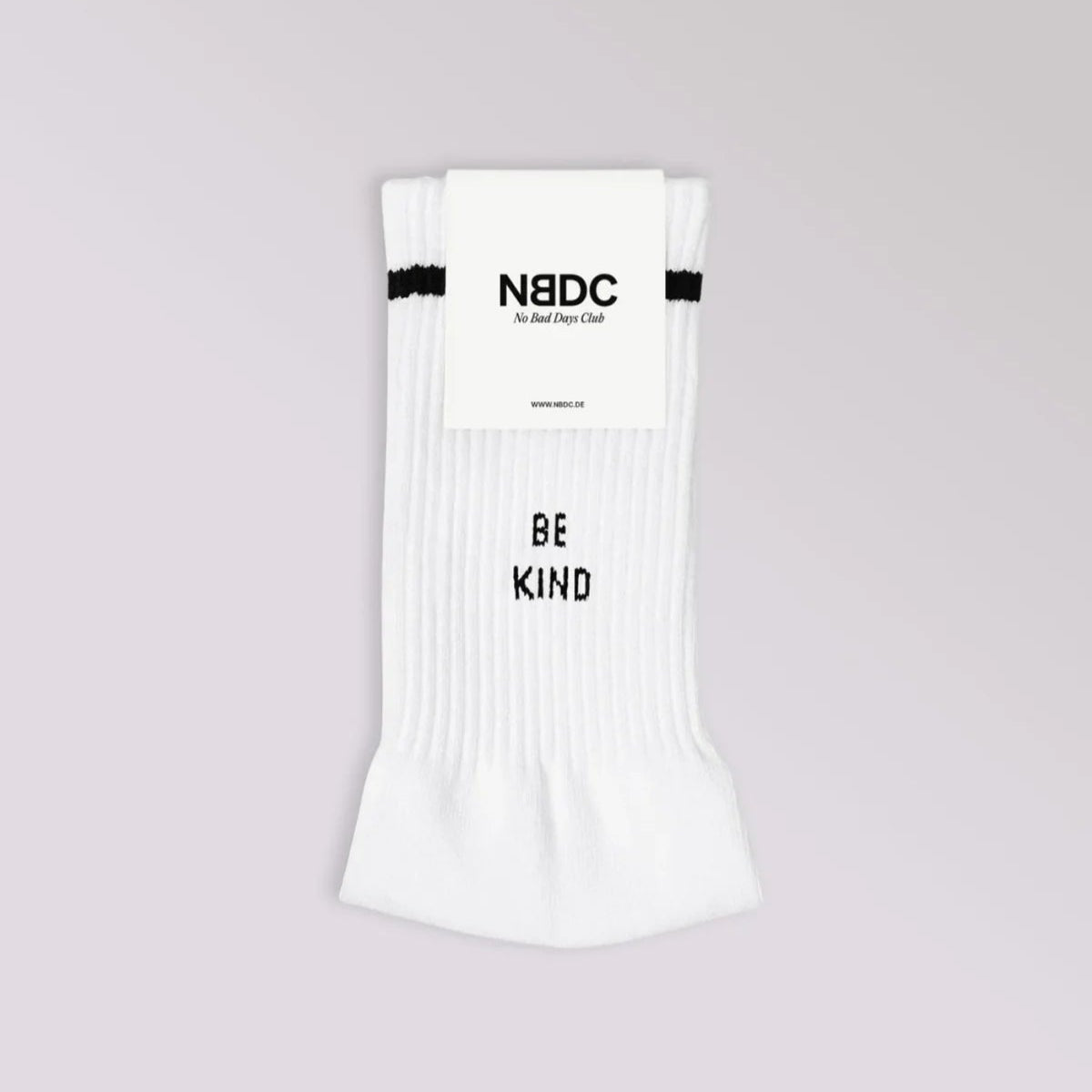 Ein Paar weiße Socken mit einem schwarzen Streifen am oberen Ende, verpackt mit einem Etikett mit der Aufschrift NBDC. Auf den Socken steht in schwarzen Großbuchstaben das Wort BE KIND. Der Hintergrund ist hellgrau.