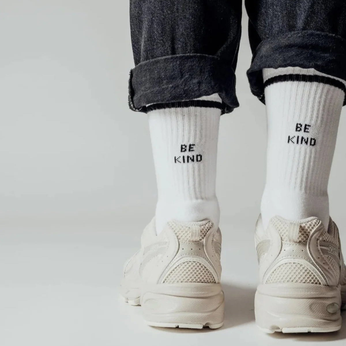 Eine Person in hochgekrempelten dunklen Jeans, weißen Turnschuhen und weißen Socken mit der Aufschrift BE KIND auf der Rückseite, die auf einer hellen Fläche steht.