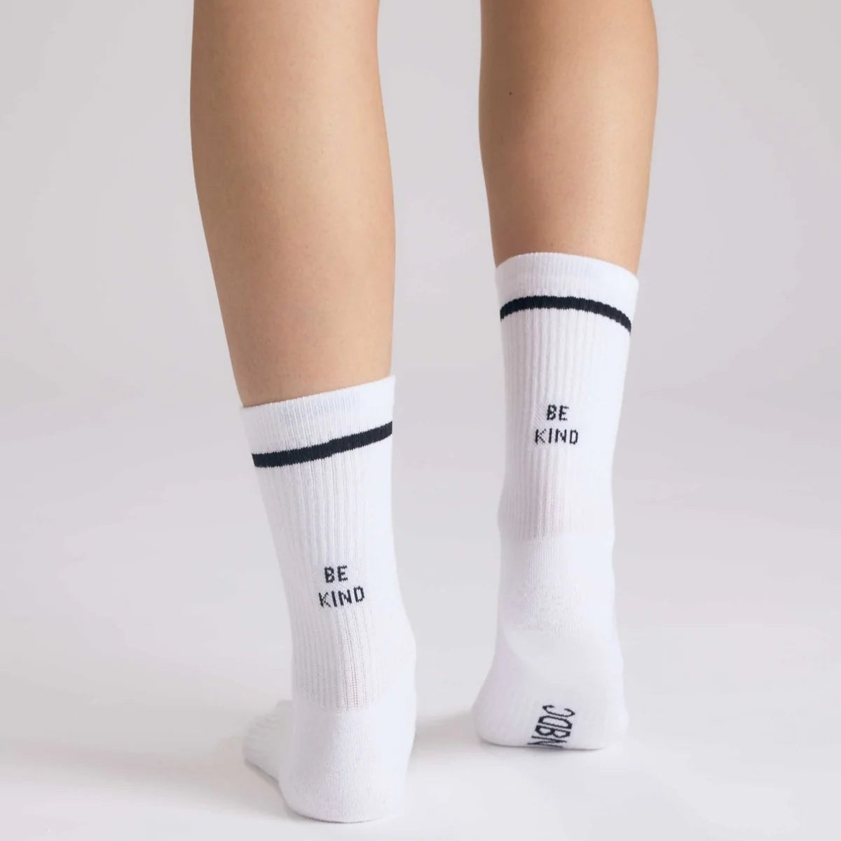 Eine Person steht auf einer weißen Fläche und trägt weiße Socken mit einem schwarzen Streifen am oberen Ende und der Aufschrift BE KIND auf der Rückseite jeder Socke.