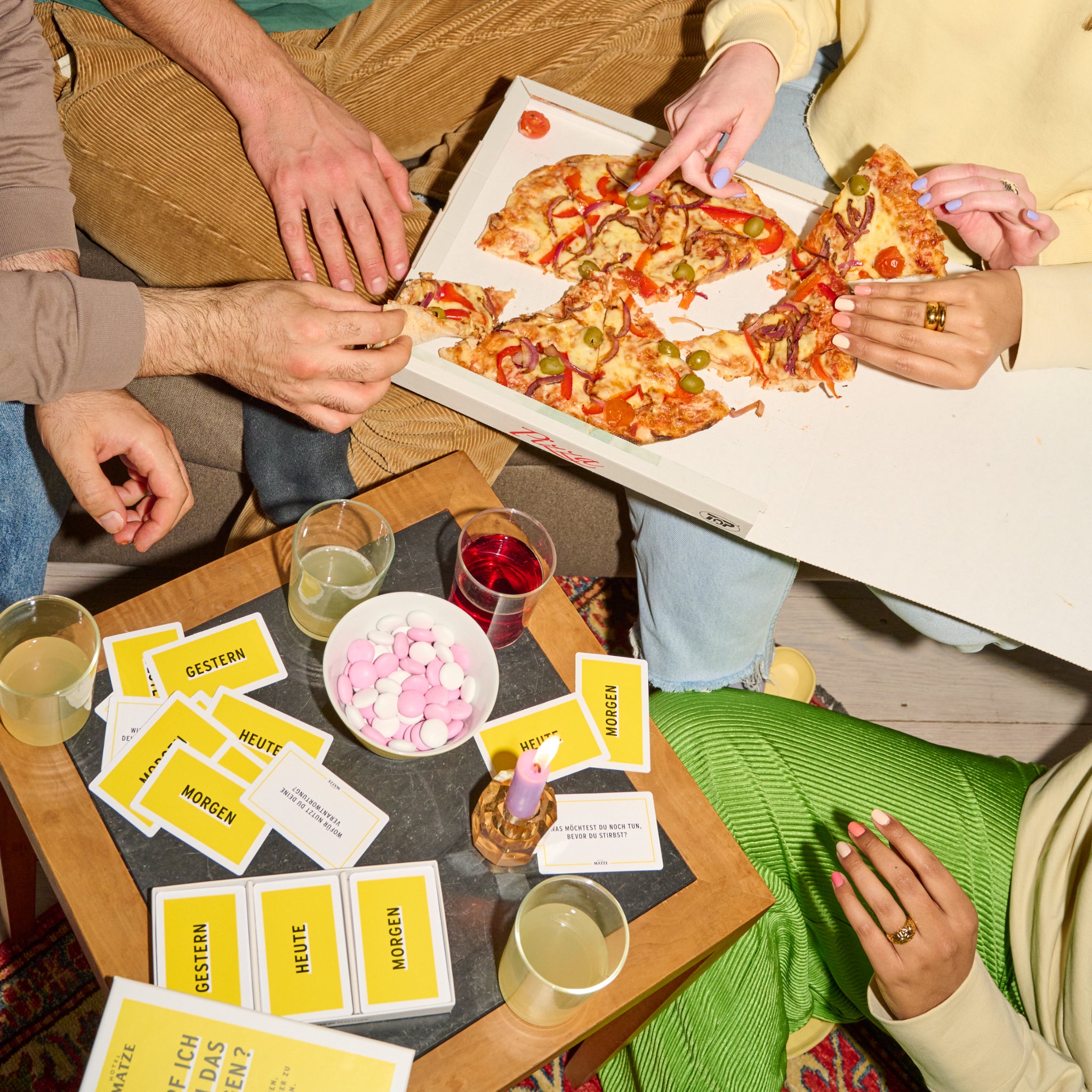 Vier Personen teilen sich eine Pizza aus einer offenen Schachtel und greifen mit ihren Händen hinein. Auf einem kleinen Tisch stehen Getränke, eine Kerze, Süßigkeiten und gelbe Karten mit deutschen Wörtern wie HEUTE und MORGEN. Es sind nur Hände und teilweise Arme zu sehen.