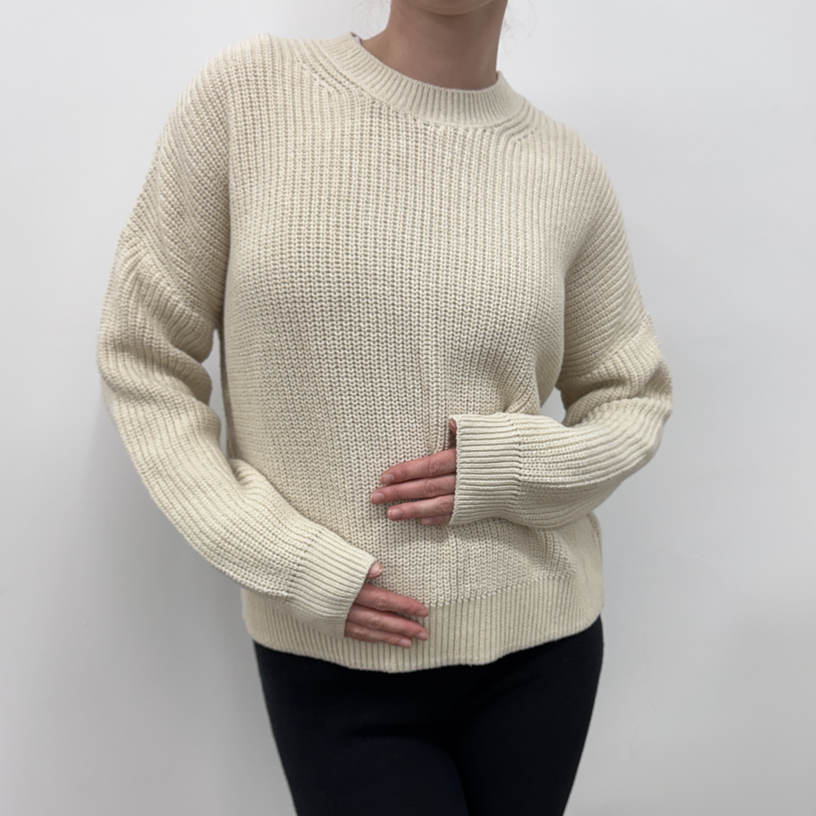 Eine Person, die einen cremefarbenen Strickpullover und eine schwarze Hose trägt, steht vor einem schlichten weißen Hintergrund und hat eine Hand auf den Bauch und die andere auf die Hüfte gestützt.
