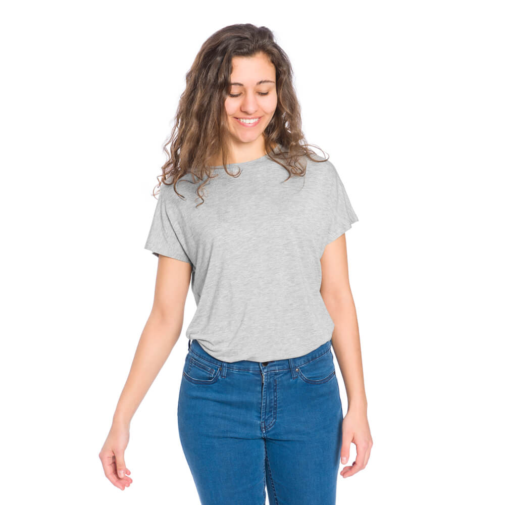 Frau mit lockigem Haar trägt ein graues T-Shirt und blaue Jeans, lächelt und schaut nach unten, vor weißem Hintergrund.