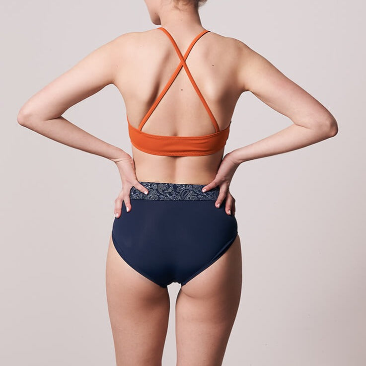 Person in orange sports bra and navy blue high-waisted shorts with hands on hips, seen from the back.