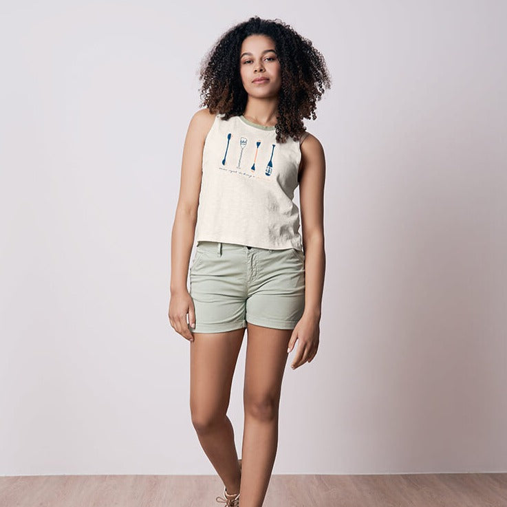 Eine Frau in einem weißen ärmellosen Top und hellgrünen Shorts steht vor einem einfachen Hintergrund.
