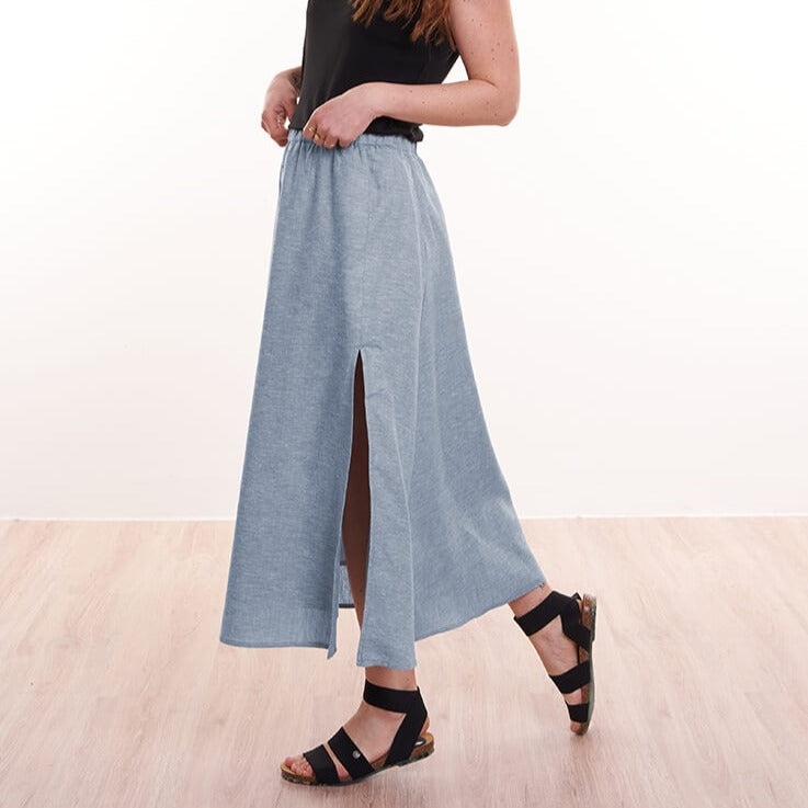 Frau in schwarzem Top und blauem Rock mit Seitenschlitz, trägt schwarze Sandalen auf hellem Holzboden.