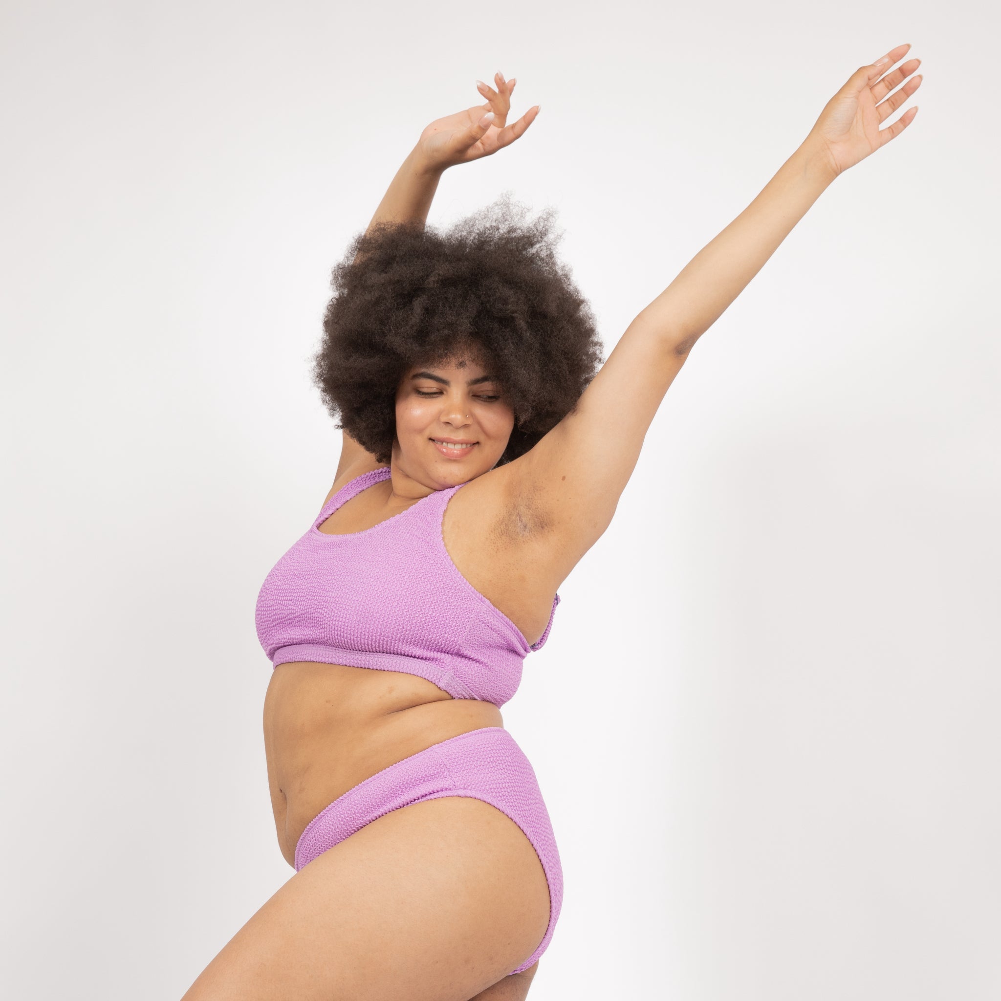 Person in lilac bikini poses joyfully with arms raised against a plain white background.