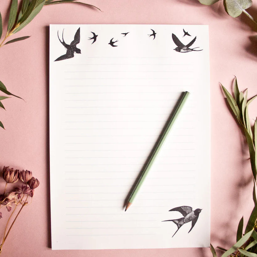 Ein Blatt liniertes Papier mit Vogeldarstellungen liegt auf einer rosa Fläche, umgeben von grünen Eukalyptuszweigen und getrockneten Blumen. Ein grüner Bleistift liegt auf dem Papier.