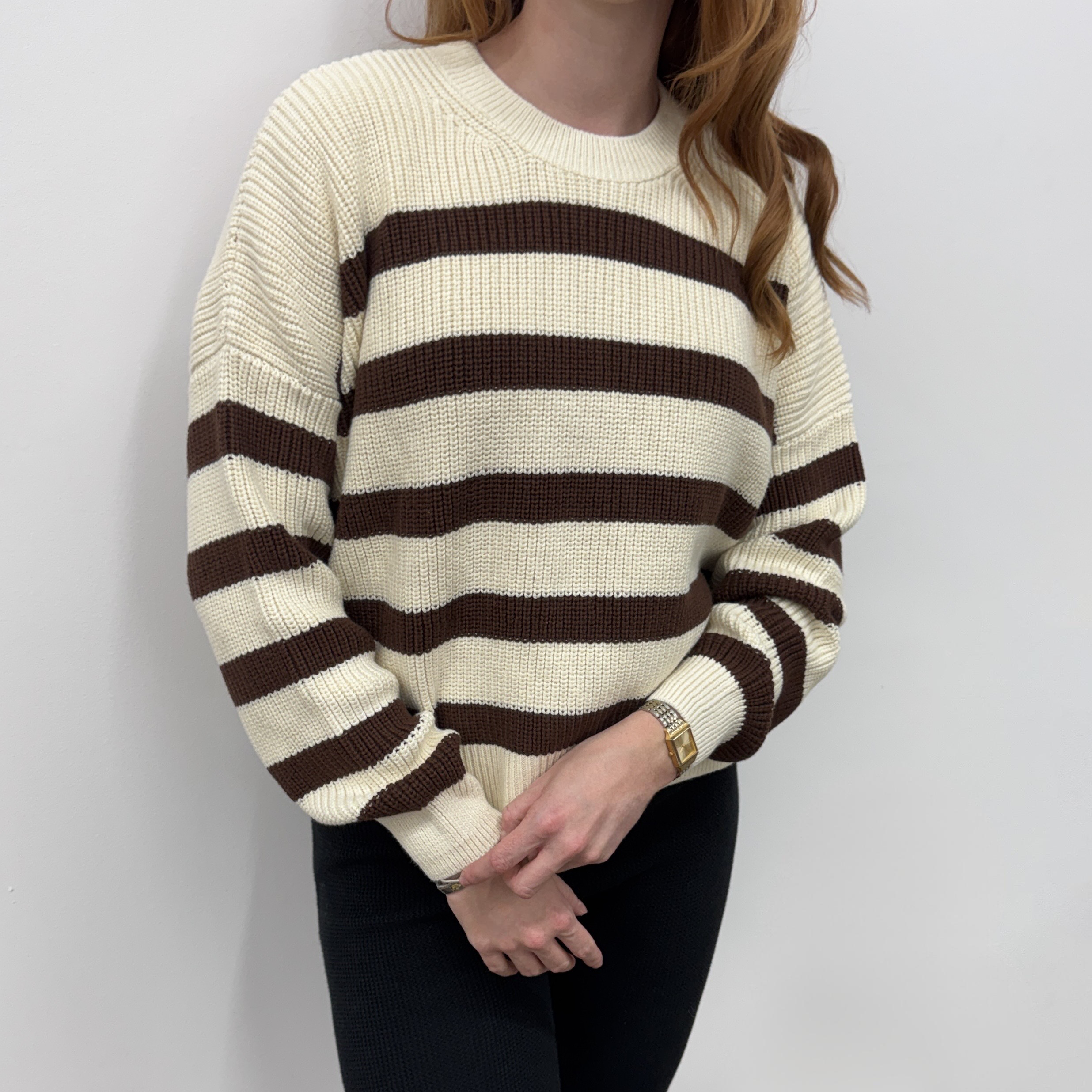 Eine Frau mit langem, gewelltem Haar trägt einen cremefarbenen Pullover mit breiten braunen Querstreifen und eine schwarze Hose vor einem schlichten weißen Hintergrund.