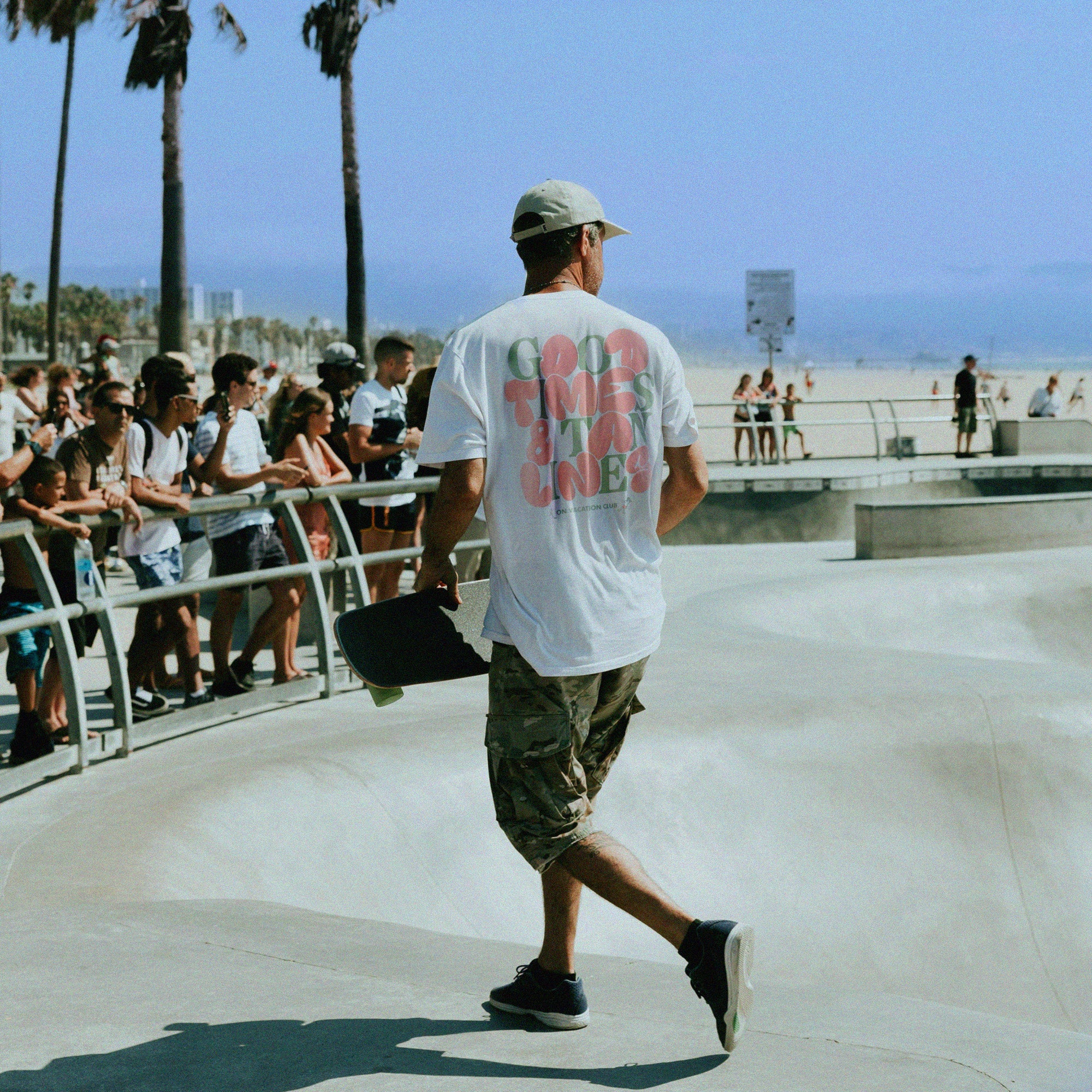 Ein Mann mit Mütze, weißem T-Shirt und Camouflage-Shorts trägt ein Skateboard in einem Skatepark am Strand. Eine Menschenmenge schaut hinter einem Geländer zu, mit Palmen und blauem Himmel im Hintergrund.