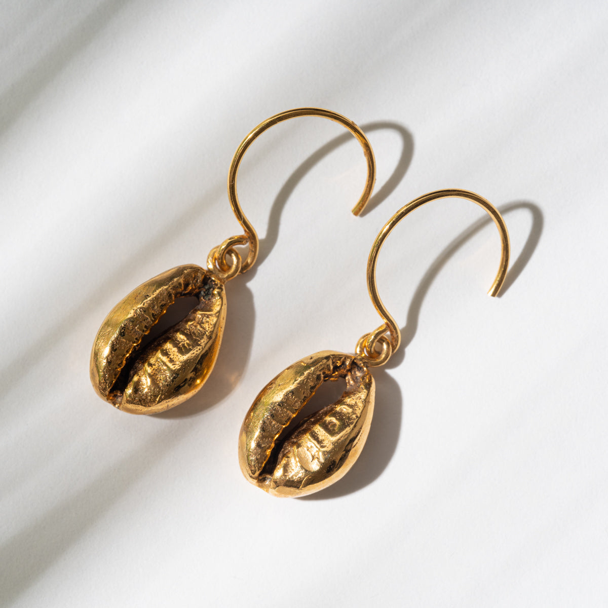 Goldene Hakenohrringe mit Anhängern in Form von Kaurimuscheln, die auf einer weißen Oberfläche mit sanften Schatten dargestellt sind.