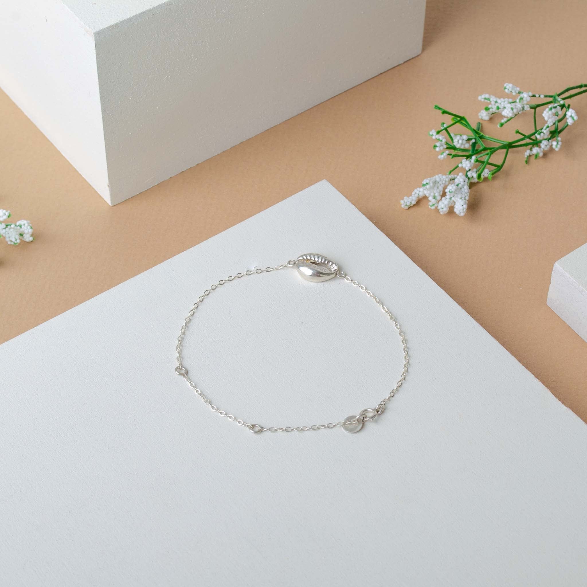Ein zartes silbernes Kettenarmband mit einem kleinen Muschelanhänger ist auf einer weißen Fläche ausgestellt, umgeben von minimalen weißen Blöcken und Zweigen mit kleinen weißen Blumen auf beigem Hintergrund.