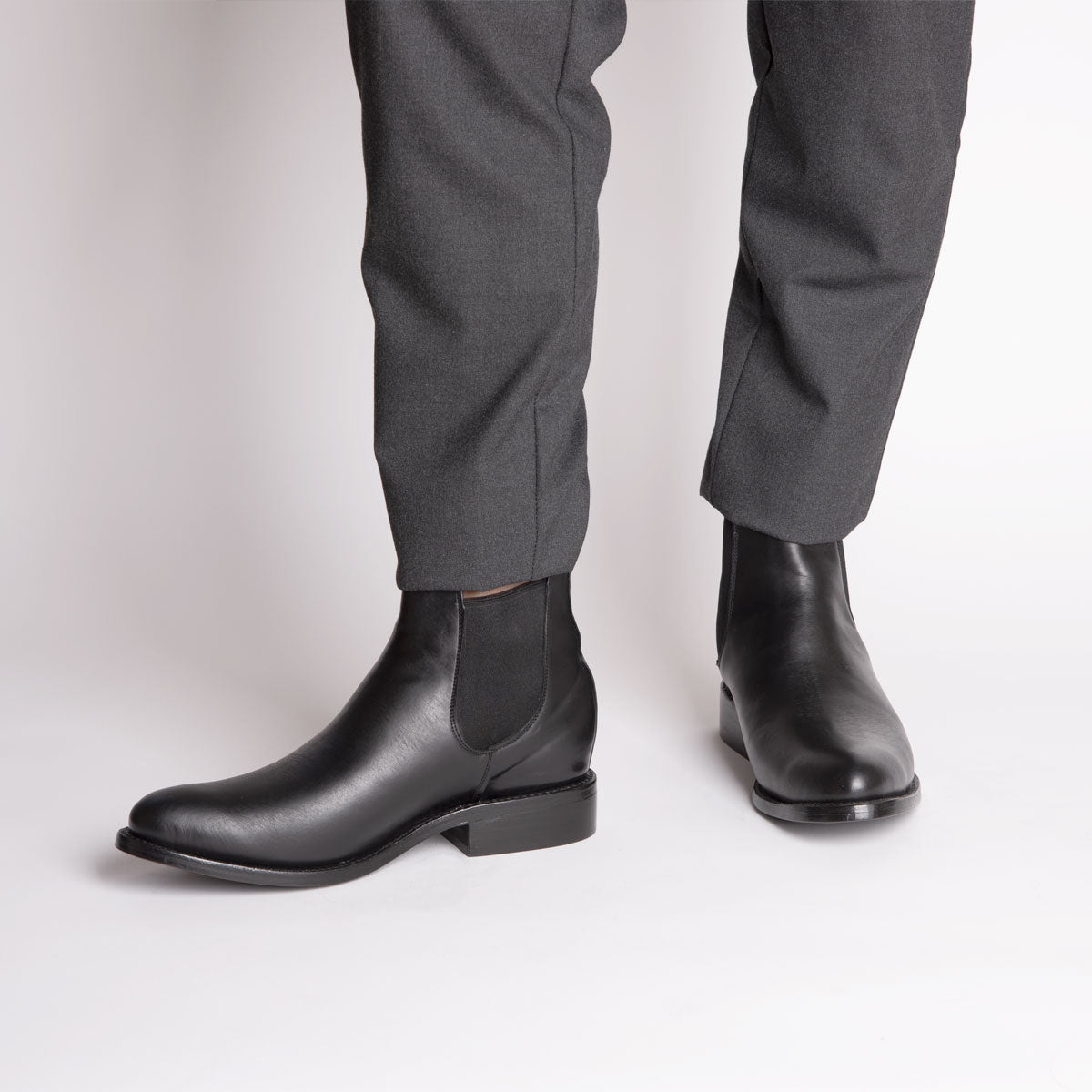 Eine Person, die eine dunkelgraue Anzughose und schwarze Chelsea-Stiefel aus Leder trägt, steht vor einem weißen Hintergrund.