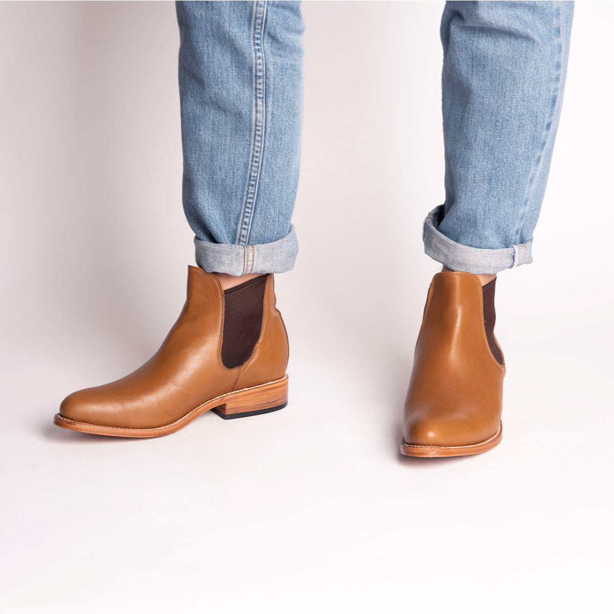 Person in hellblauen, hochgekrempelten Jeans und hellbraunen Lederstiefeln mit dunklen, elastischen Seiteneinsätzen, stehend vor einem schlichten weißen Hintergrund.