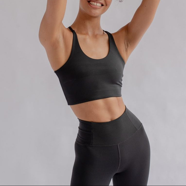 Eine lächelnde Person mit lockigem Haar posiert mit den Händen in den Haaren, trägt einen schwarzen Sport-BH und schwarze Leggings mit hoher Taille vor einem hellen Hintergrund.