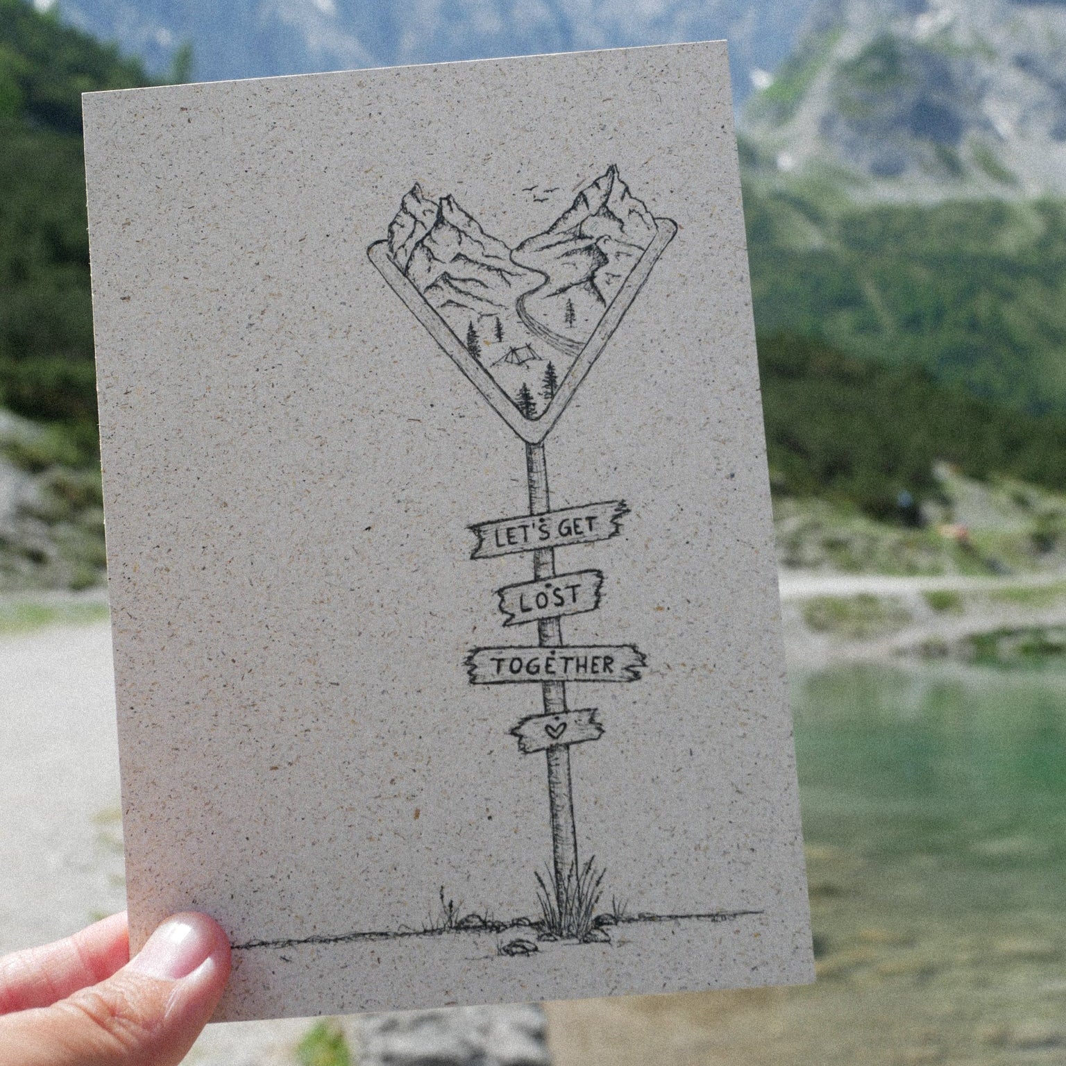 Eine Hand hält eine Postkarte mit einer Zeichnung von Bergen, einem Fluss und Schildern mit der Aufschrift Lets get lost together vor einer malerischen Bergsee-Landschaft.