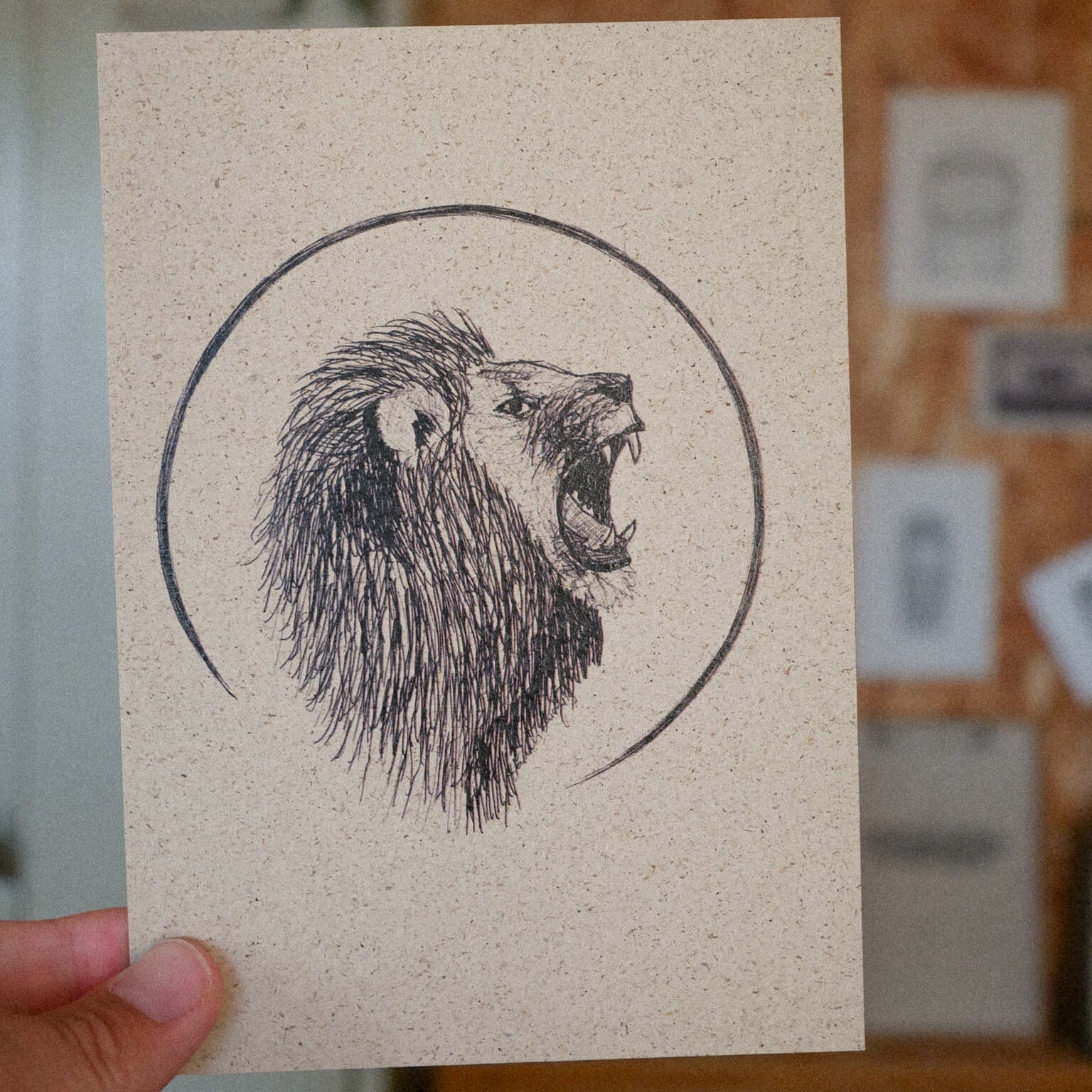 Eine Hand hält eine Karte mit einer schwarzen Tuschezeichnung eines brüllenden Löwenkopfes innerhalb eines Kreises. Der Hintergrund ist ein Innenraum, in dem unscharfe Wandkunst und eine Pflanze zu sehen sind.