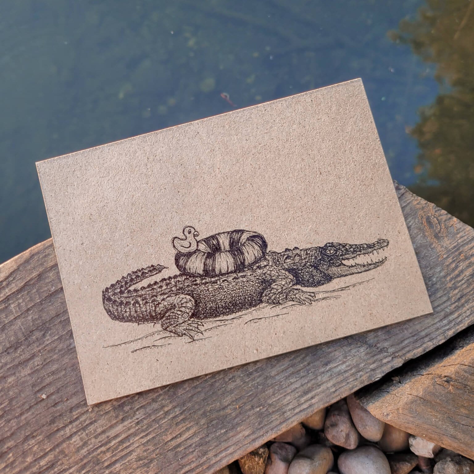 Eine Postkarte mit einer schwarzen Skizze eines Krokodils, das einen Kürbis und eine kleine Ente auf dem Rücken trägt, liegt auf einem Holzbrett in der Nähe eines Teiches, wobei im Hintergrund Kieselsteine und Wasser zu sehen sind.