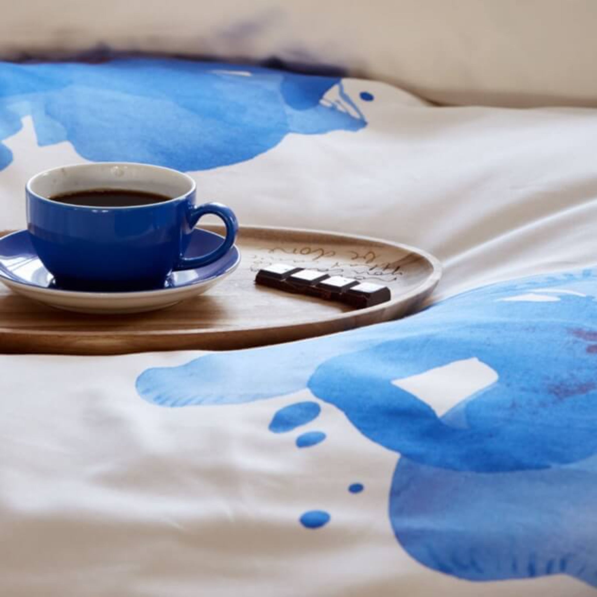 Blaue Kaffeetasse auf Holztablett mit Schokoladenstücken, auf einem Bett mit blauer Musterbettwäsche.