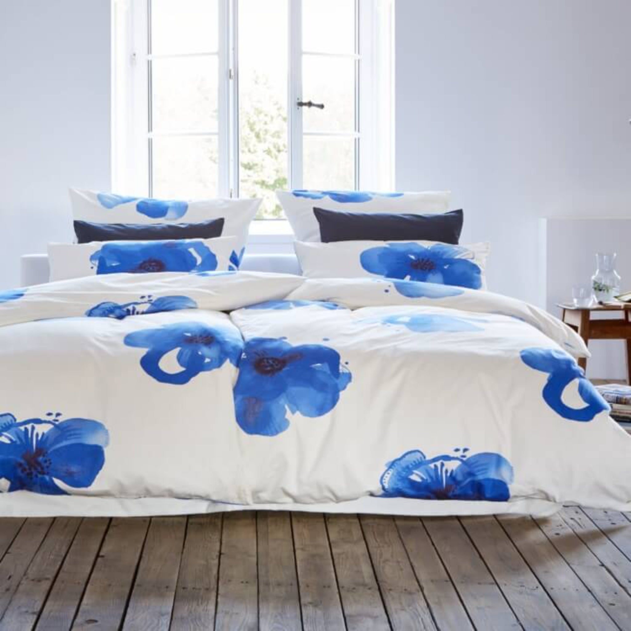 Bett mit weißer Bettwäsche und blauen Blumenmotiven vor einem Fenster auf Holzboden.