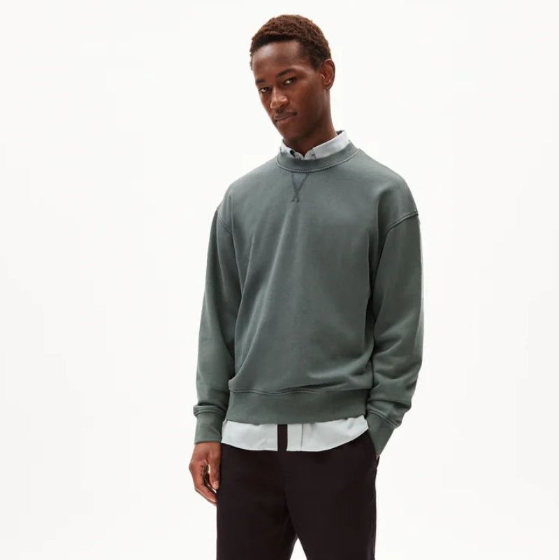 Ein Mann steht vor einem weißen Hintergrund und trägt ein gedämpftes grünes Sweatshirt über einem hellen Hemd mit Kragen, eine dunkle Hose und beigefarbene Pantoffelschuhe. Er hat die Hände in den Taschen und einen entspannten Gesichtsausdruck.