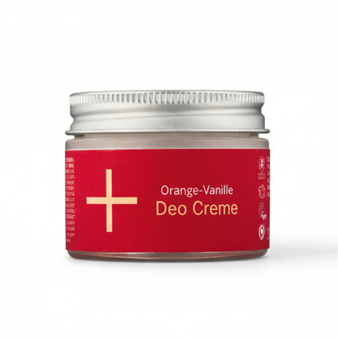 Ein kleiner Glastiegel mit einem silbernen Deckel, der Orange-Vanille Deo Creme enthält. Der Tiegel hat ein rotes Etikett mit weißem Text und einem weißen Pluszeichen auf der linken Seite. Der Hintergrund ist weiß.