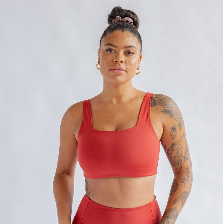 Eine Frau mit Tattoos, die einen roten Sport-BH und passende Leggings trägt, steht selbstbewusst vor einem hellgrauen Hintergrund und blickt direkt in die Kamera, wobei sie ihr Haar zu einem Dutt gebunden hat.