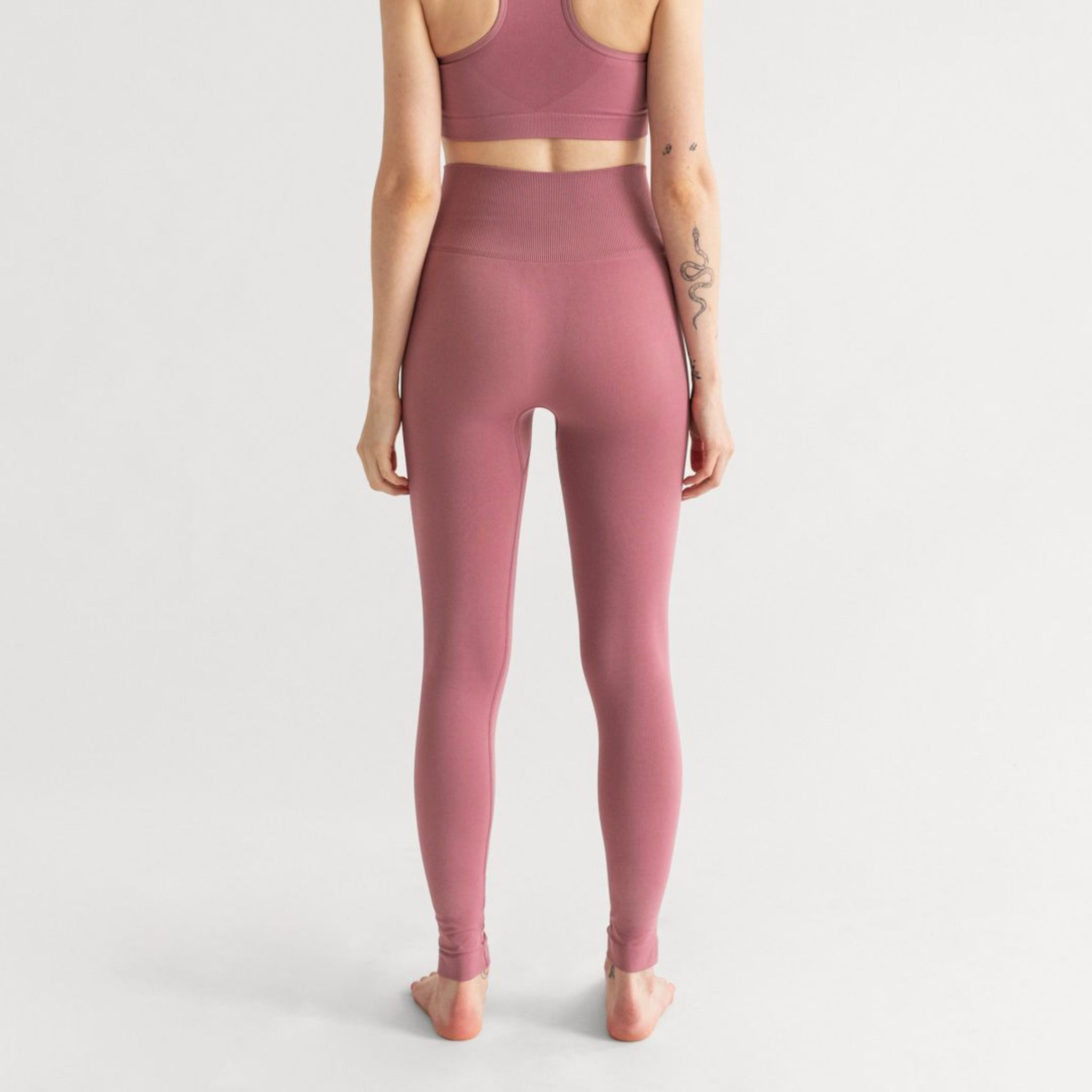 Person in rosafarbenem Sport-BH und Leggings steht barfuß mit Rücken zur Kamera, mit Tattoos auf dem Arm.