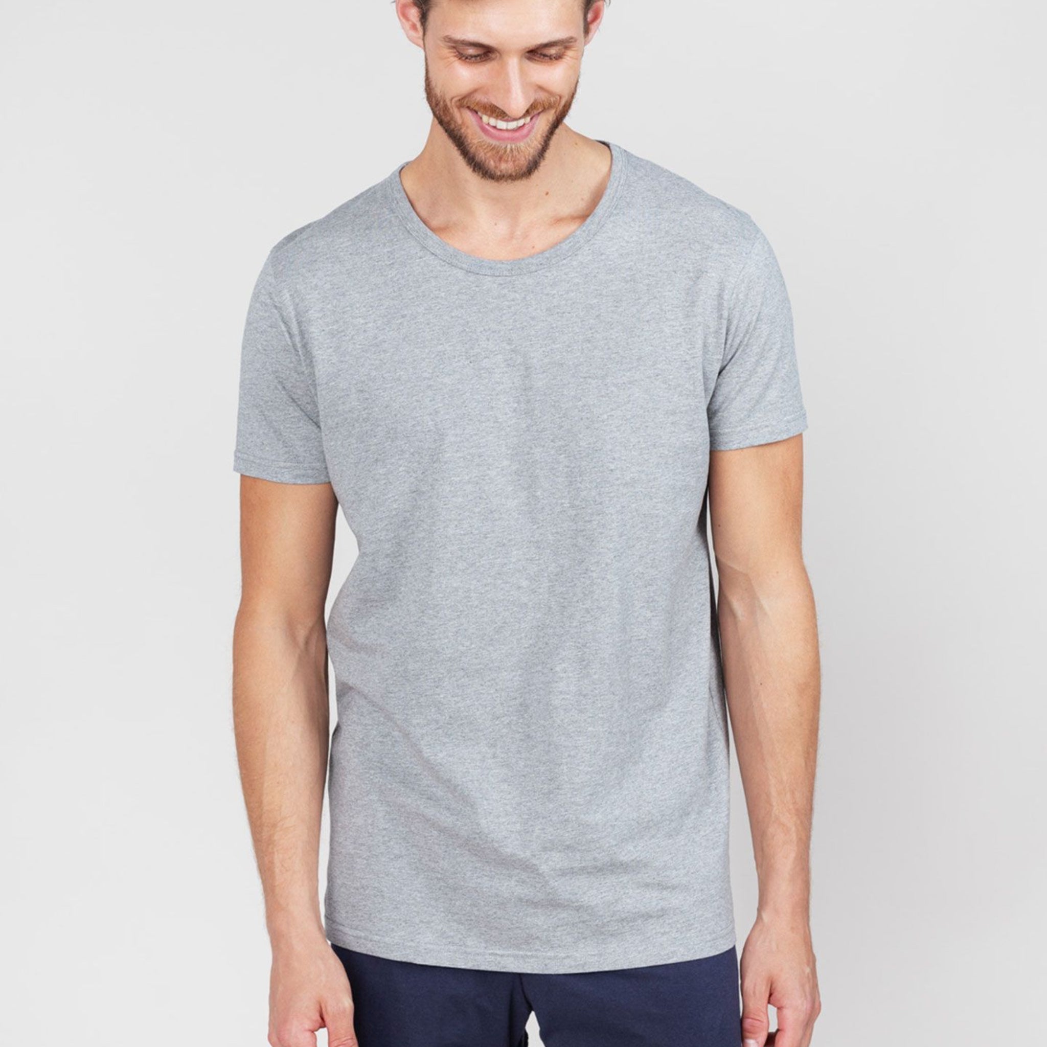 Ein Mann trägt ein graues T-Shirt und dunkle Hosen, lächelt und schaut nach unten vor einem hellen Hintergrund.