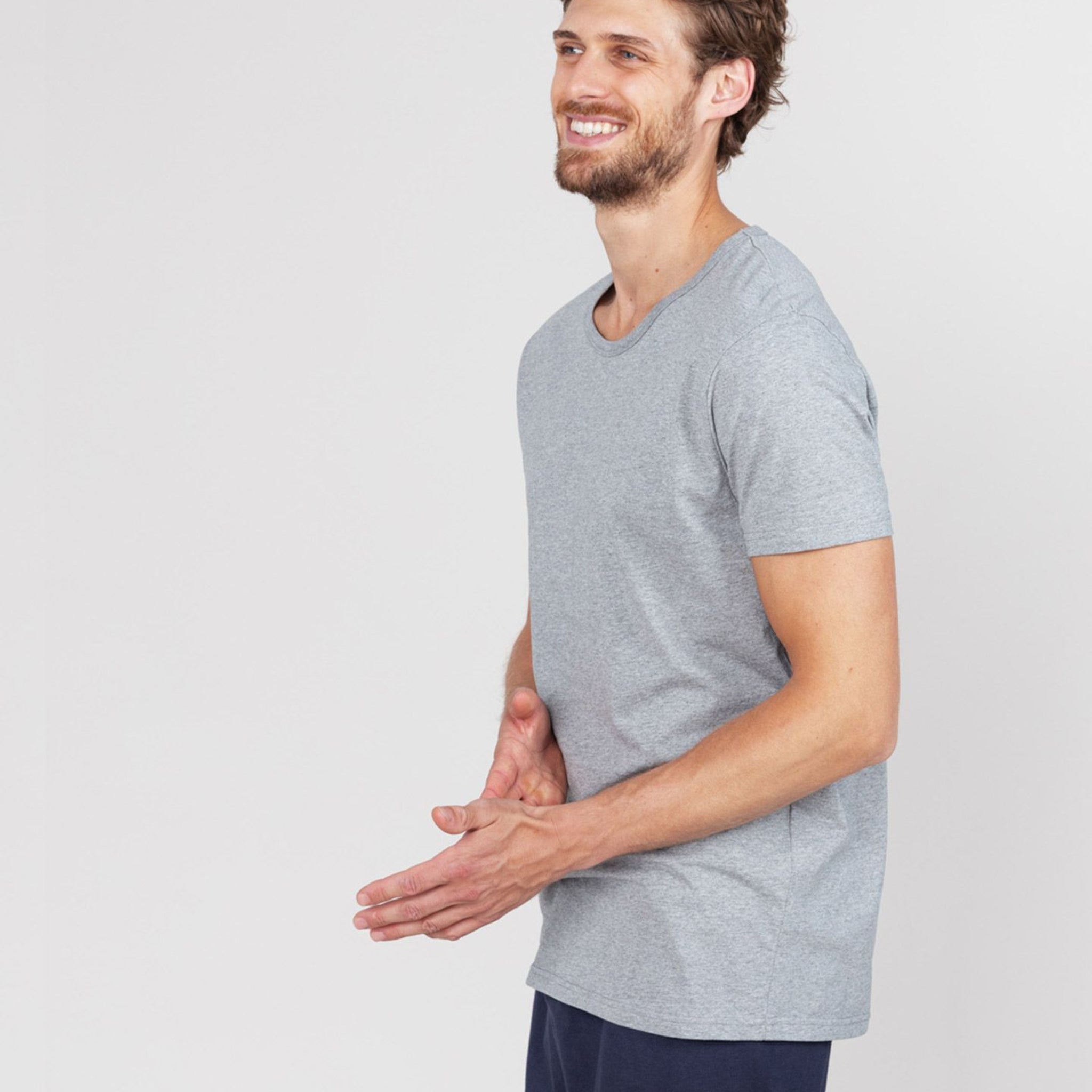 Mann in grauem T-Shirt und dunklen Hose lächelt und schaut zur Seite, Hände entspannt vor sich.
