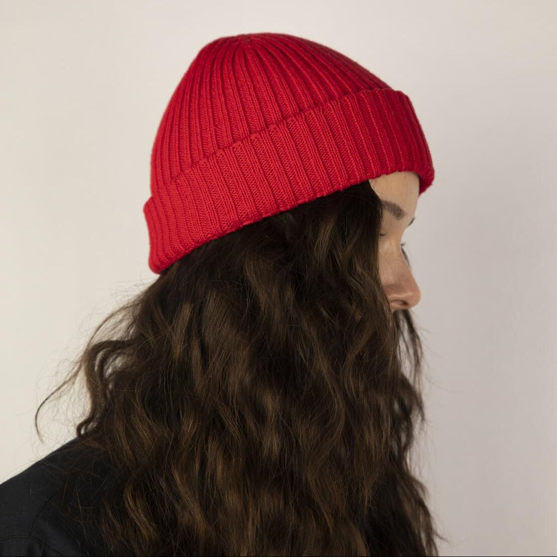 Eine Person mit langen, gewellten braunen Haaren, die eine leuchtend rote Strickmütze trägt, wird im Profil vor einem schlichten hellen Hintergrund gezeigt.