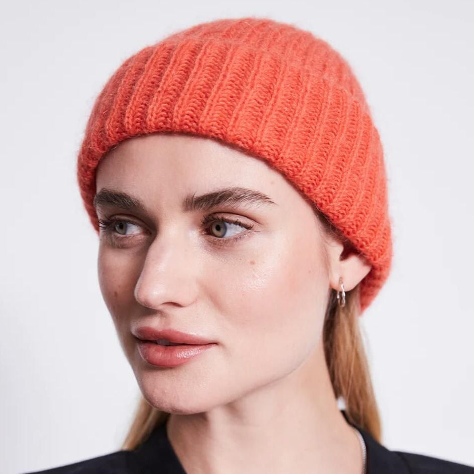 Eine Frau mit heller Haut und hellbraunem Haar trägt eine leuchtend orangefarbene Strickmütze und ein dunkles Oberteil. Sie blickt leicht zur Seite und hat einen neutralen Gesichtsausdruck vor einem schlichten hellen Hintergrund.