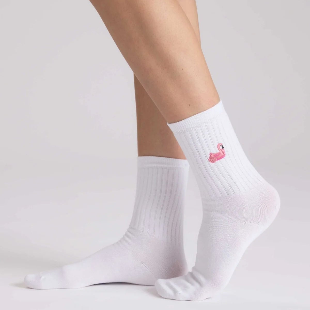 Eine Person, die weiße gerippte Socken trägt, auf die ein kleiner rosa Schwan gestickt ist, steht vor einem hellen Hintergrund.