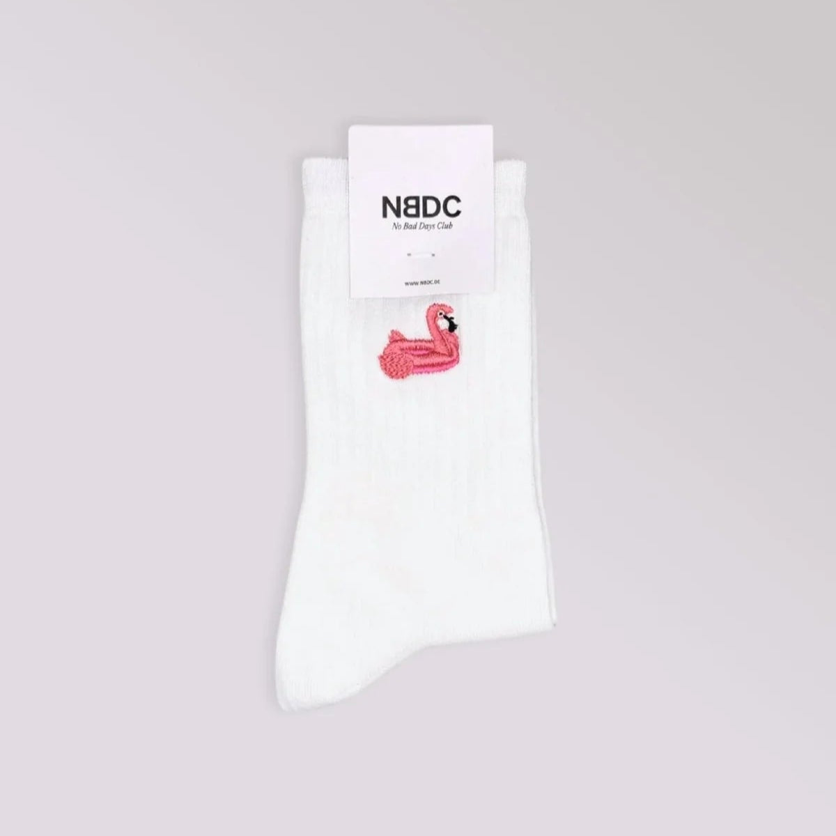 Ein Paar weiße Socken mit einem kleinen gestickten rosa Flamingo vor einem hellgrauen Hintergrund. Oben ist ein Etikett mit N8DC und zusätzlichem Text angebracht.