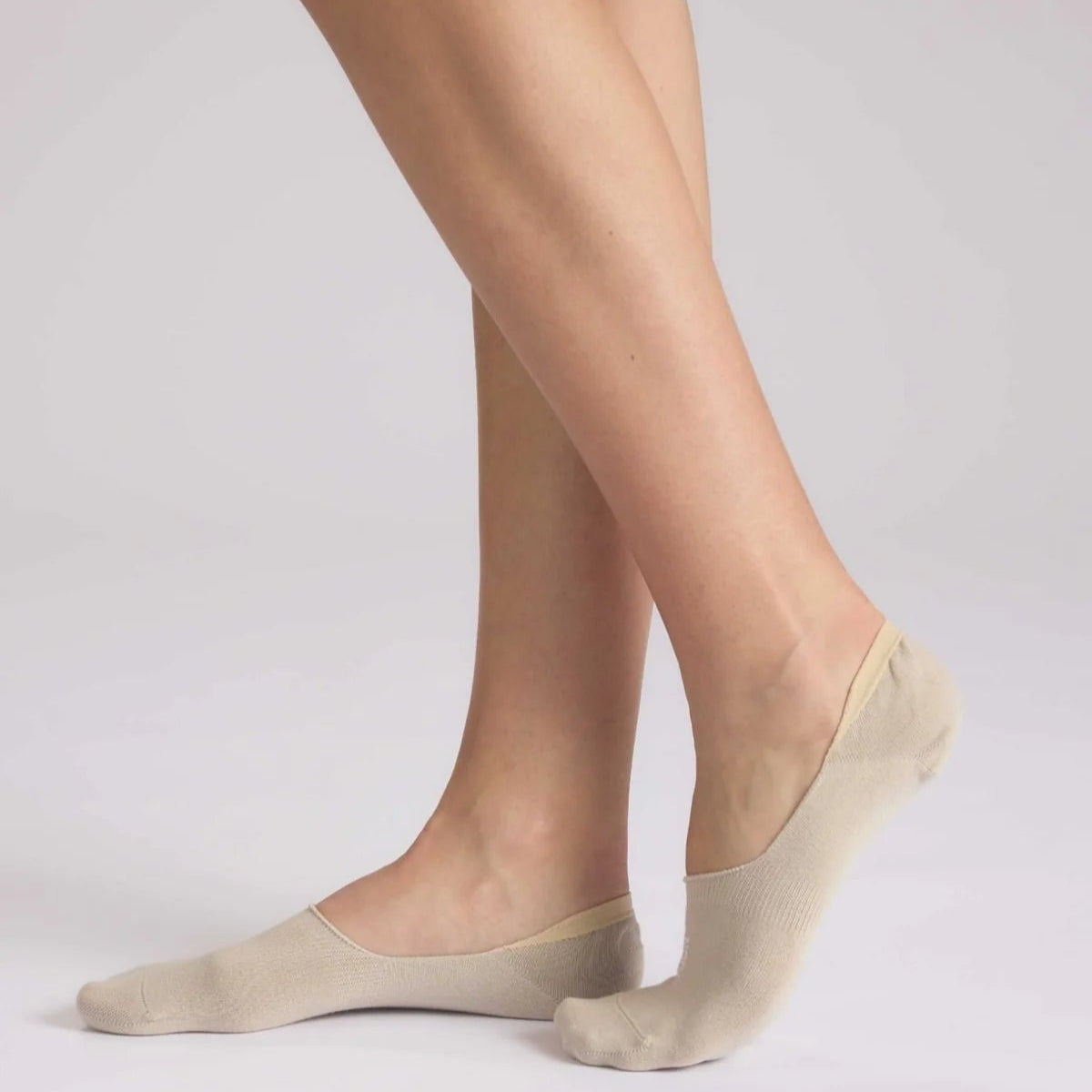 Eine Person, die beigefarbene No-Show-Socken trägt, steht auf Zehenspitzen und hat ein Bein leicht vor dem anderen gekreuzt, vor einem schlichten hellen Hintergrund.