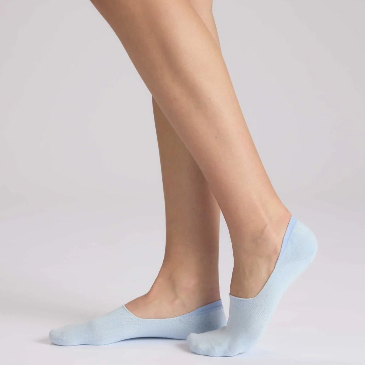 Eine Person, die hellblaue Socken trägt, steht auf einer weißen Fläche, hat einen Fuß leicht angehoben und die Beine an den Knöcheln gekreuzt.