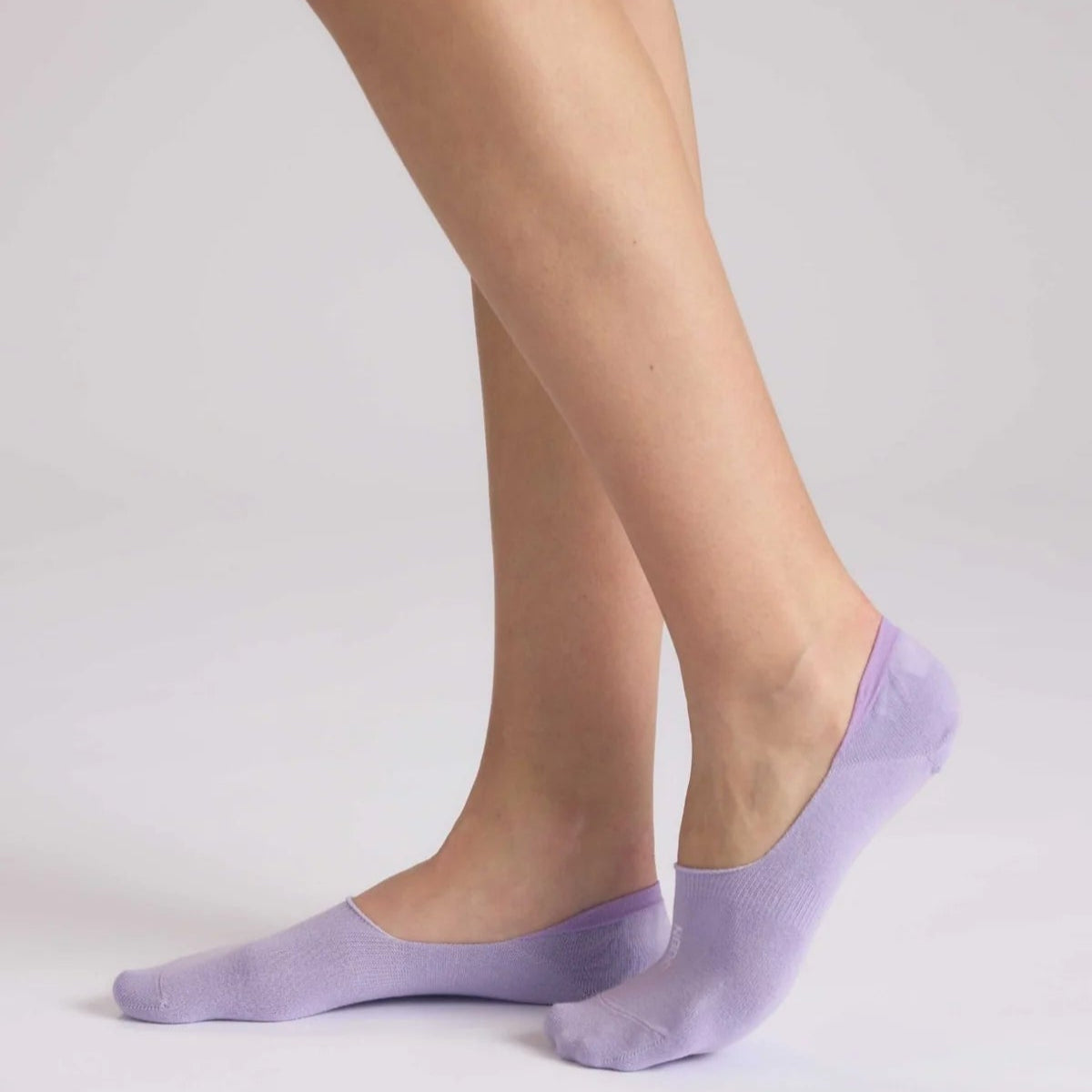 Eine Person, die auf Zehenspitzen steht und lavendelfarbene No-Show-Socken vor einem schlichten, hellen Hintergrund trägt.