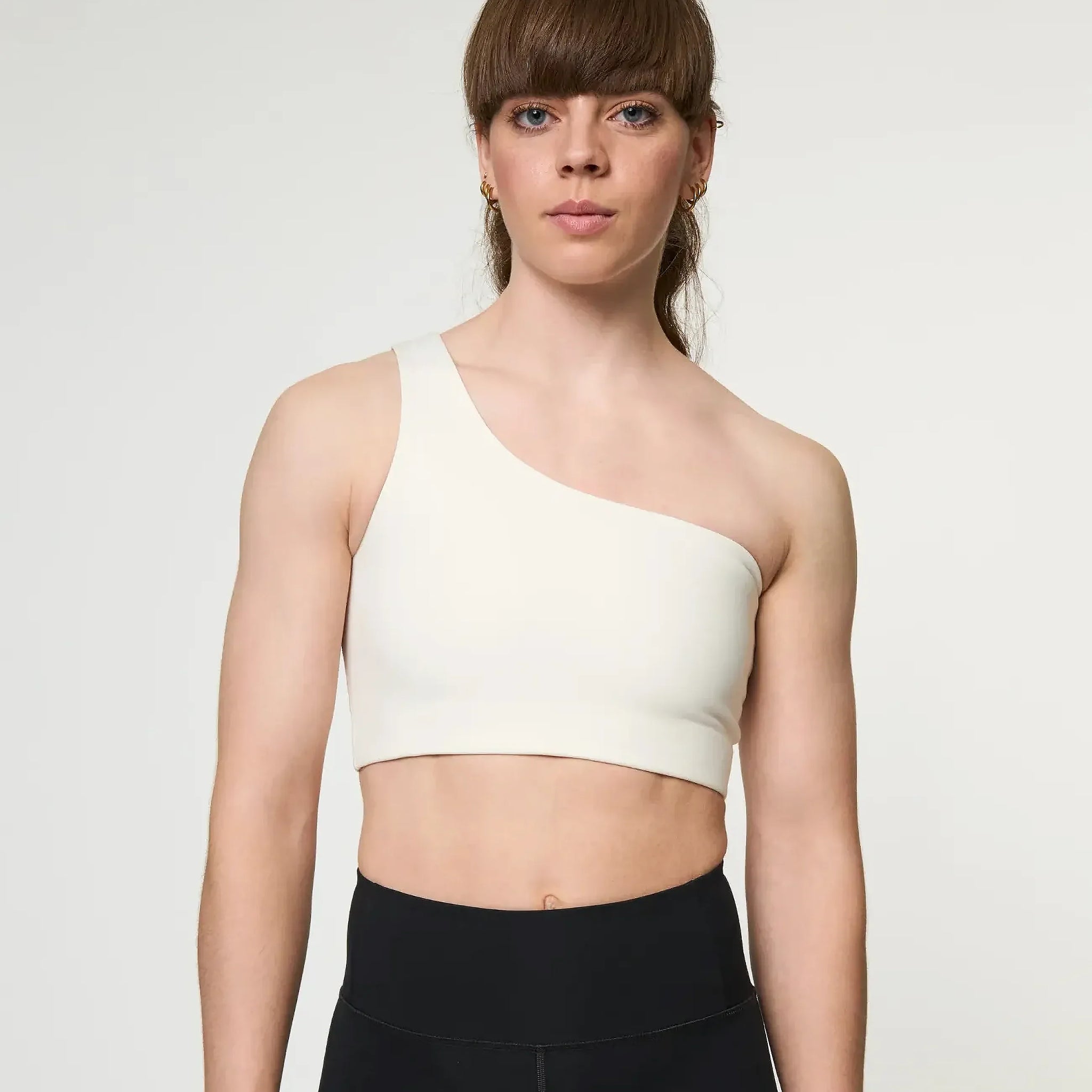 Eine Frau mit braunem Haar und Pony trägt einen cremefarbenen, schulterfreien Sport-BH und schwarze Leggings mit hoher Taille und steht vor einem schlichten, hellen Hintergrund.