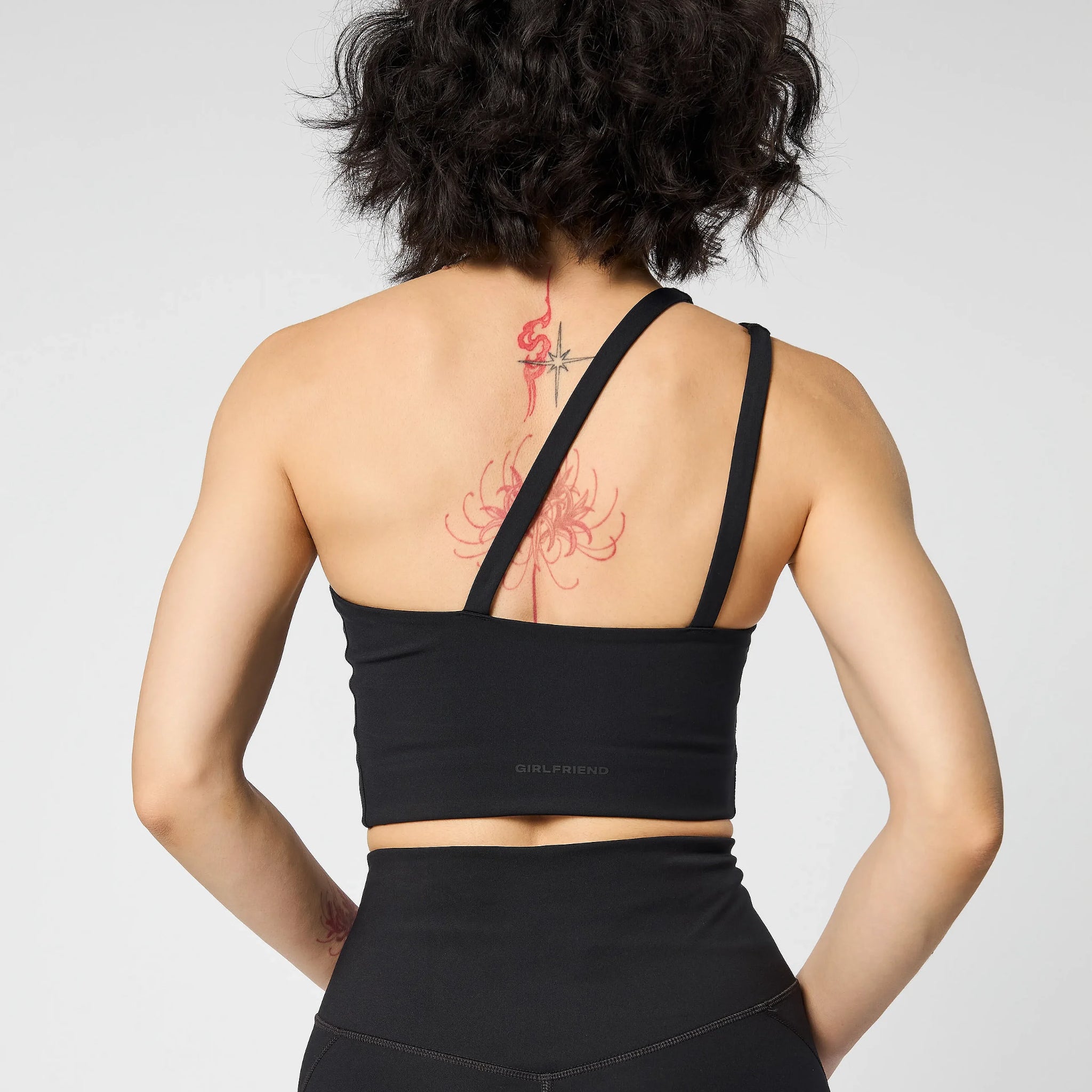 Eine Person mit kurzen, gewellten dunklen Haaren ist von hinten zu sehen. Sie trägt ein schwarzes, schulterfreies Crop-Top und hoch sitzende Leggings, auf denen rote Tätowierungen auf dem oberen Rücken und der Wirbelsäule zu sehen sind.