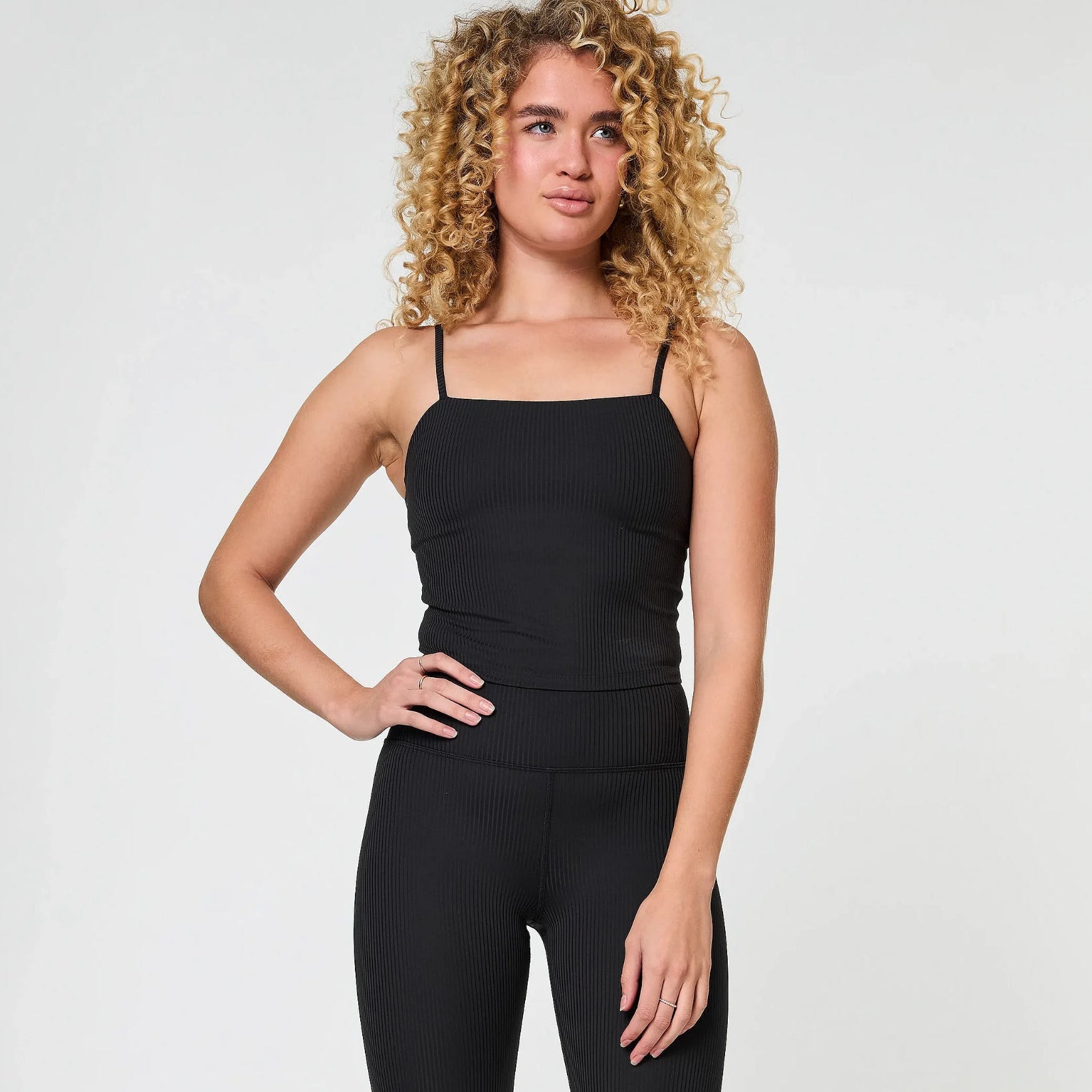 Eine Frau mit lockigem blondem Haar trägt ein schwarzes geripptes Oberteil und dazu passende Leggings mit hoher Taille. Sie steht vor einem schlichten hellgrauen Hintergrund und hat eine Hand an der Hüfte.