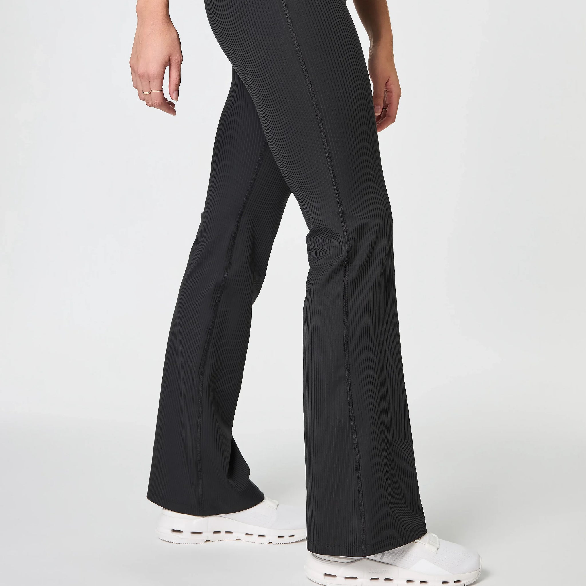 Eine Person in schwarzen Schlaghosen und weißen Turnschuhen steht vor einem schlichten hellgrauen Hintergrund. Nur die Beine und ein Teil der Arme sind sichtbar.