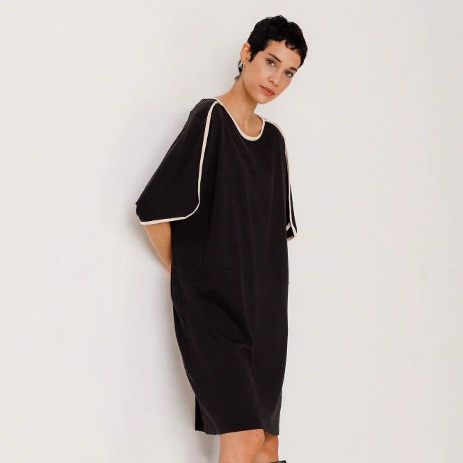 Eine Person mit kurzen dunklen Haaren trägt ein locker sitzendes schwarzes Kleid mit weißem Besatz an den Ärmeln und am Ausschnitt, gepaart mit kniehohen schwarzen Lederstiefeln, und steht vor einem schlichten weißen Hintergrund.