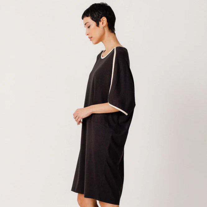 Eine Person mit kurzen dunklen Haaren, die ein lockeres schwarzes Kleid mit weißem Saum an den Ärmeln trägt, steht im Profil und hat hohe schwarze Stiefel vor einem schlichten hellen Hintergrund an.